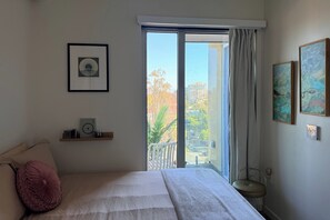 Een slaapkamer met een bed, een raam met gordijnen, een plank met spullen en ingelijste foto's aan de muur.