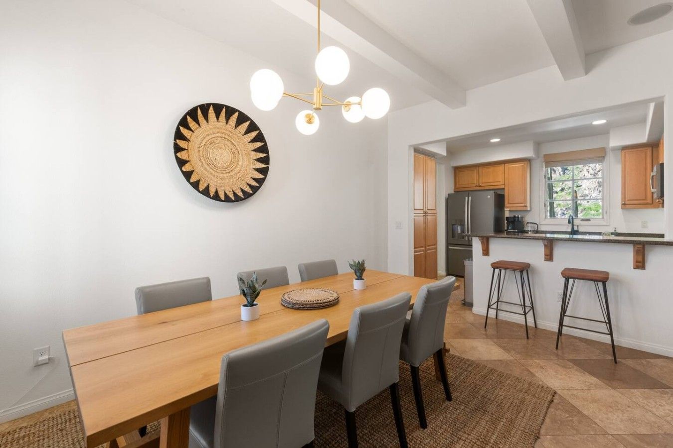A modern dining area with a wooden table, grey chairs, a decorative wall piece, and a kitchen with wooden cabinets and a refrigerator.