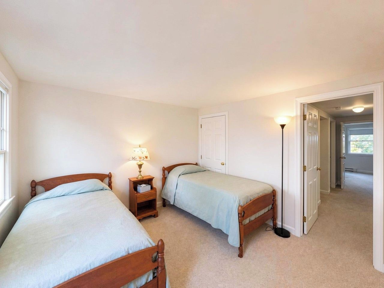 A bedroom with two single beds, a bedside table with a lamp, and a door leading to another room.