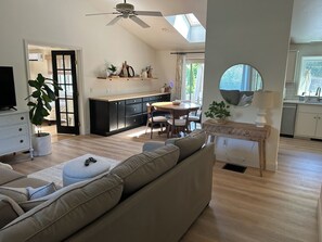A modern living room with a sofa, a dining table, and a kitchen area with black cabinets and a skylight. - Welcome to your Cape Cod getaway! ONLY 1.5 miles to Menauhant Beach, this charmi (East Falmouth)