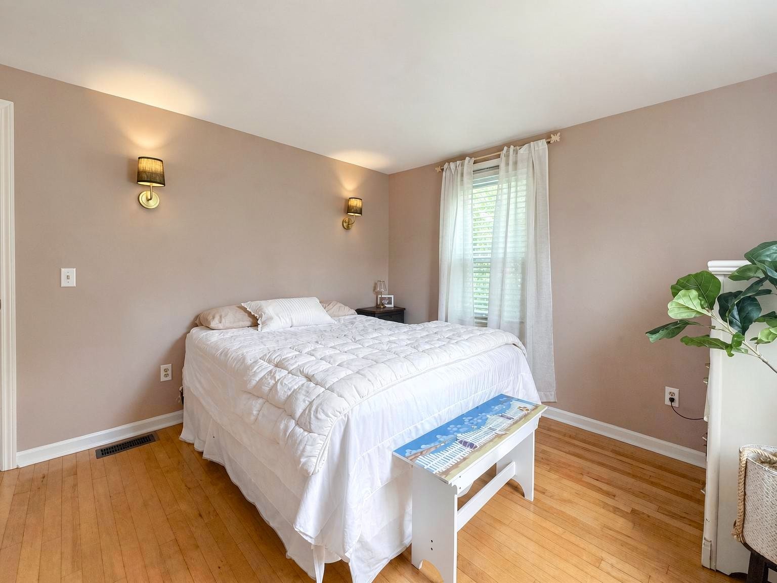 Quarto com cama, luminárias de cabeceira, janela com cortinas, planta e mesa de cabeceira.