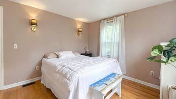 Quarto com cama, luminárias de cabeceira, janela com cortinas, planta e mesa de cabeceira.