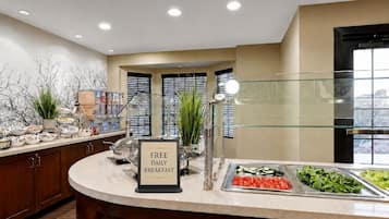 Een buffetruimte met een bord waarop staat aangegeven gratis dagelijks ontbijt, diverse voedselopties en een modern interieur.