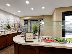 A buffet area with a sign indicating free daily breakfast, a variety of food options, and a modern interior design.