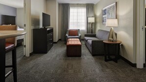 A hotel room with a flat-screen TV, a sofa, a chair, a small table with a lamp, and a window with curtains.