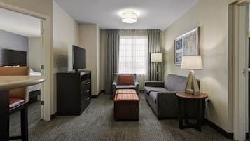 Een hotelkamer met een flatscreen televisie, een bank, een stoel, een klein tafeltje met een lamp en een raam met gordijnen.