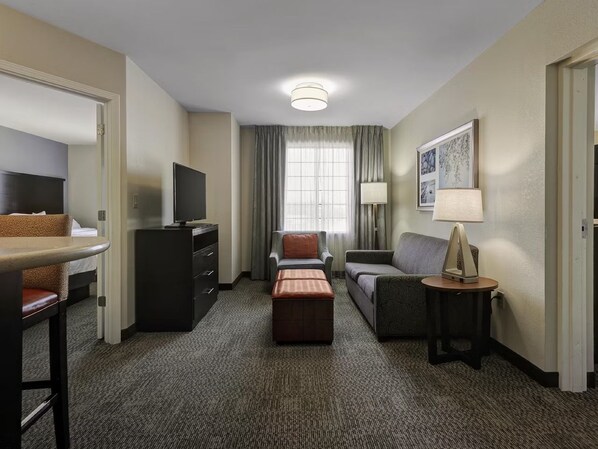 Une chambre d’hôtel équipée d’un téléviseur à écran plat, d’un canapé, d’un fauteuil, d’une petite table avec une lampe et d’une fenêtre avec des rideaux.