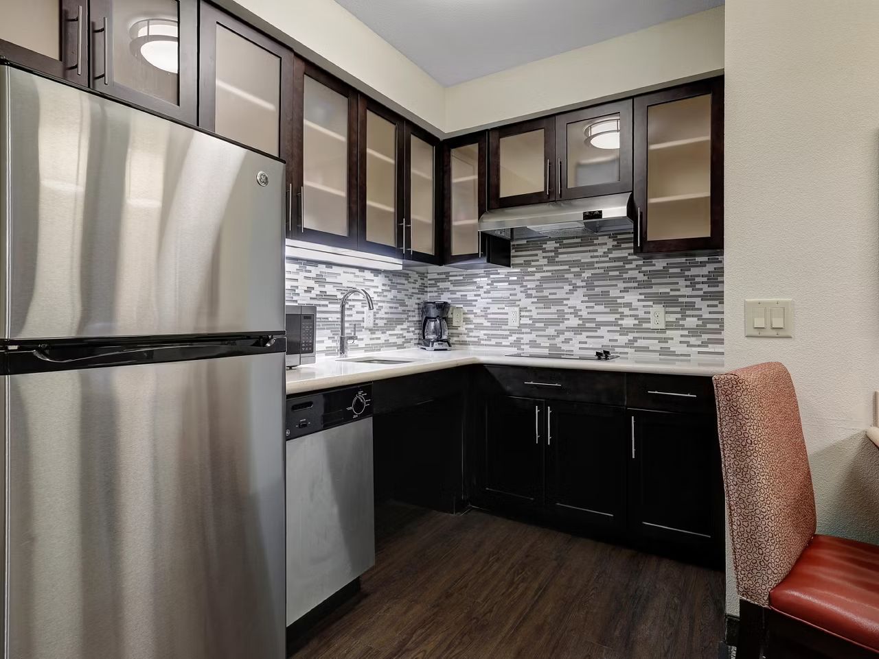 A compact kitchen with a stainless steel refrigerator, dark wooden cabinets, a tiled backsplash, and a red upholstered chair.