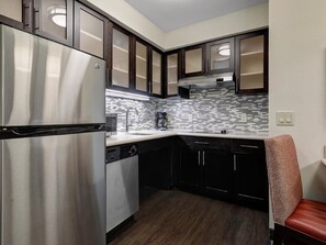 A compact kitchen with a stainless steel refrigerator, dark wooden cabinets, a tiled backsplash, and a red upholstered chair.