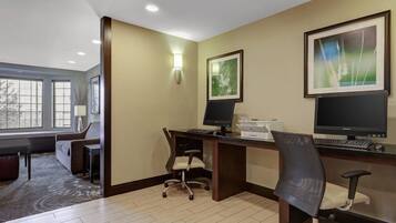 A hotel room with a desk, two chairs, a computer, and a large window.