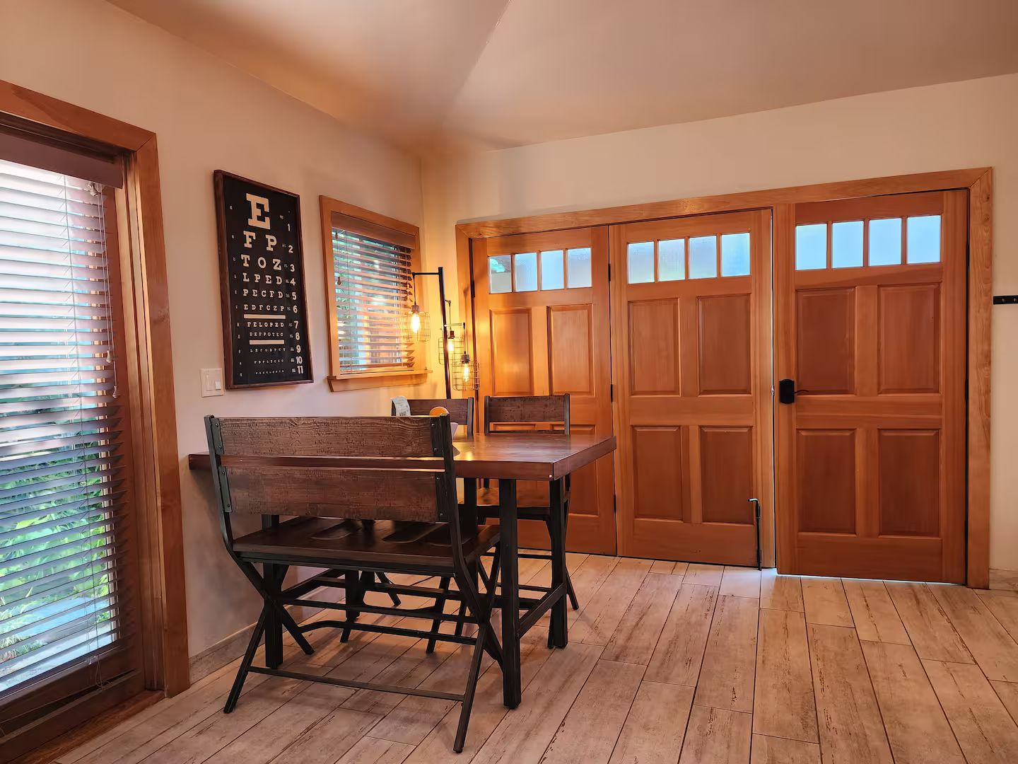 A dining area with a wooden table and chairs, a wall-mounted eye chart, and a set of wooden double doors.