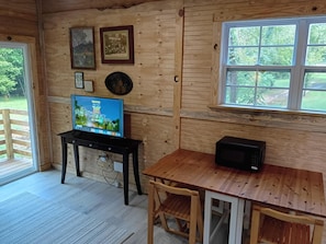 A wooden cabin interior with a television on a table, a microwave, and a window with a view of greenery.