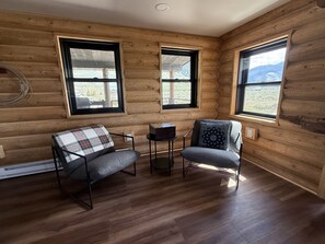 L’intérieur d’une cabane en rondins, avec deux fenêtres, une cheminée et un coin salon agrémenté d’un coussin à carreaux et d’un oreiller à motifs.
