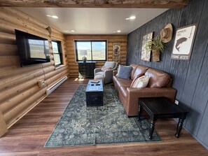 A log cabin interior with a leather sofa, a television mounted on the wall, and a large window offering a view of the outdoors.
