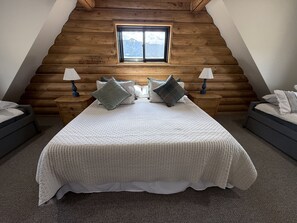 Et soveværelse med skråt loft, en stor seng med quiltet dyne, to sengeborde med lamper og et vindue med udsigt til et bjerglandskab.