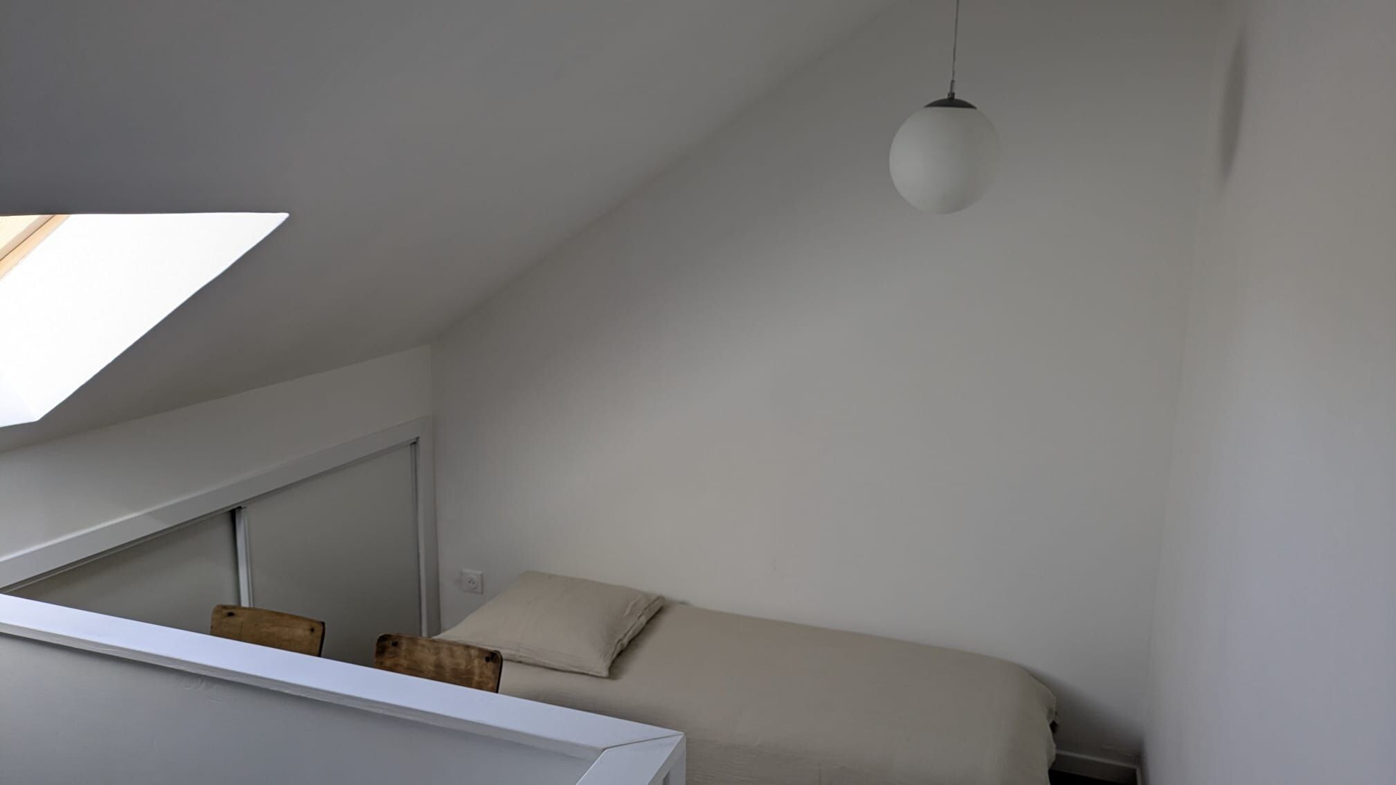 Ein schlicht eingerichtetes Schlafzimmer mit schräger Decke, einem Einzelbett, einer Hängeleuchte und einem Dachfenster.