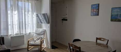 A living room with a TV, a dining table with chairs, a sofa, and a window with sheer curtains.