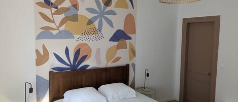 A bedroom with a bed, a wall with abstract art, a wooden door, and a hanging light fixture.