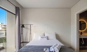 A hotel room with a bed, a bedside table, a lamp, a window with curtains, and a view of buildings outside.