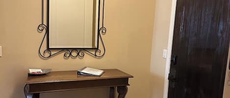 Una consola de madera oscura con un espejo encima, un pequeño adorno en el estante inferior y una puerta oscura a la derecha.
