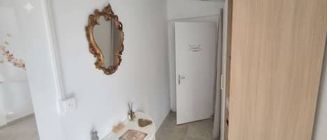 Un pasillo con una puerta blanca, un espejo y una mesita con un cuaderno y objetos decorativos.