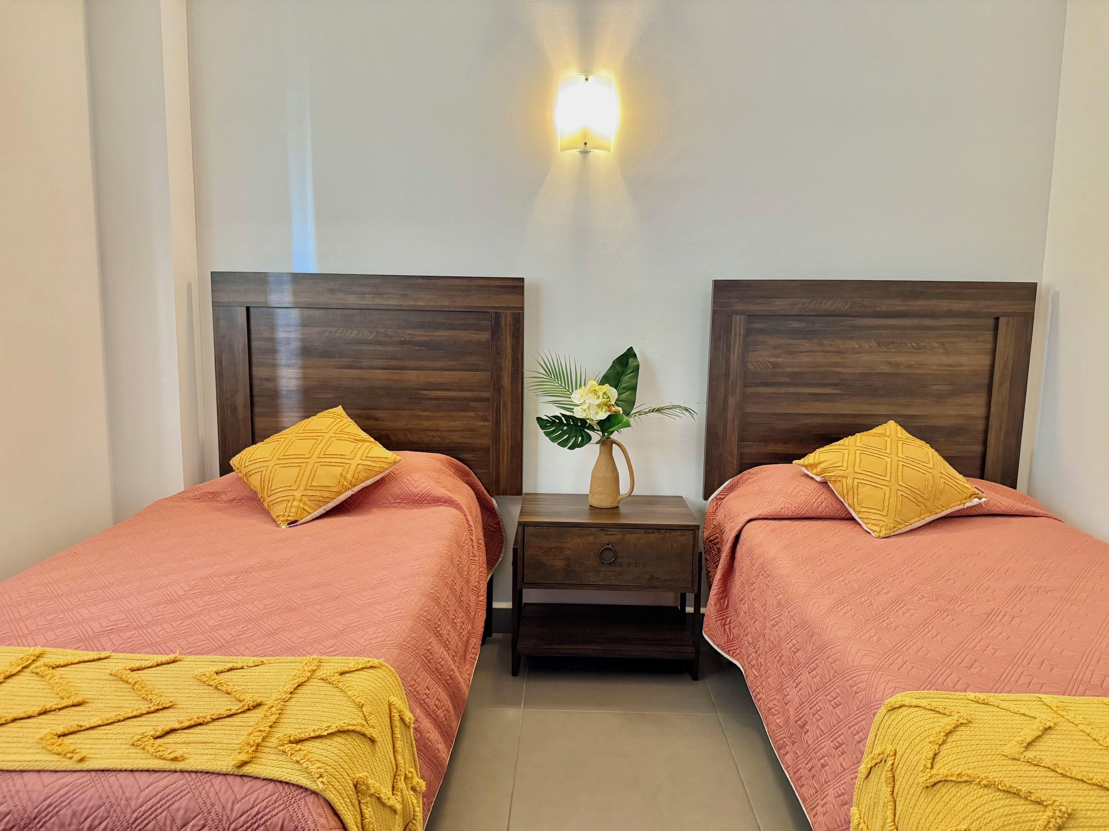 Ein Doppelzimmer mit zwei Betten, jeweils mit einem Holz-Kopfteil, einem Nachttisch und einem Beistelltisch. Über dem Nachttisch befindet sich eine Wandleuchte.