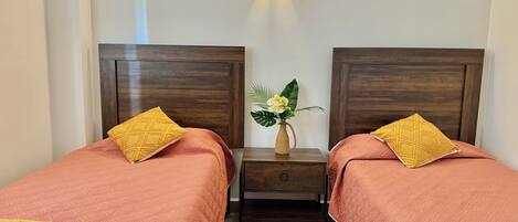 Ein Doppelzimmer mit zwei Betten, jeweils mit einem Holz-Kopfteil, einem Nachttisch und einem Beistelltisch. Über dem Nachttisch befindet sich eine Wandleuchte.