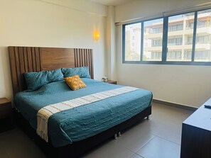 A modern bedroom with a large bed, a wooden headboard, a bedside table, and a window with a view of buildings. - Beautiful 2 Bedroom with A/C, BBQ, and Pools (Nuevo Nayarit)