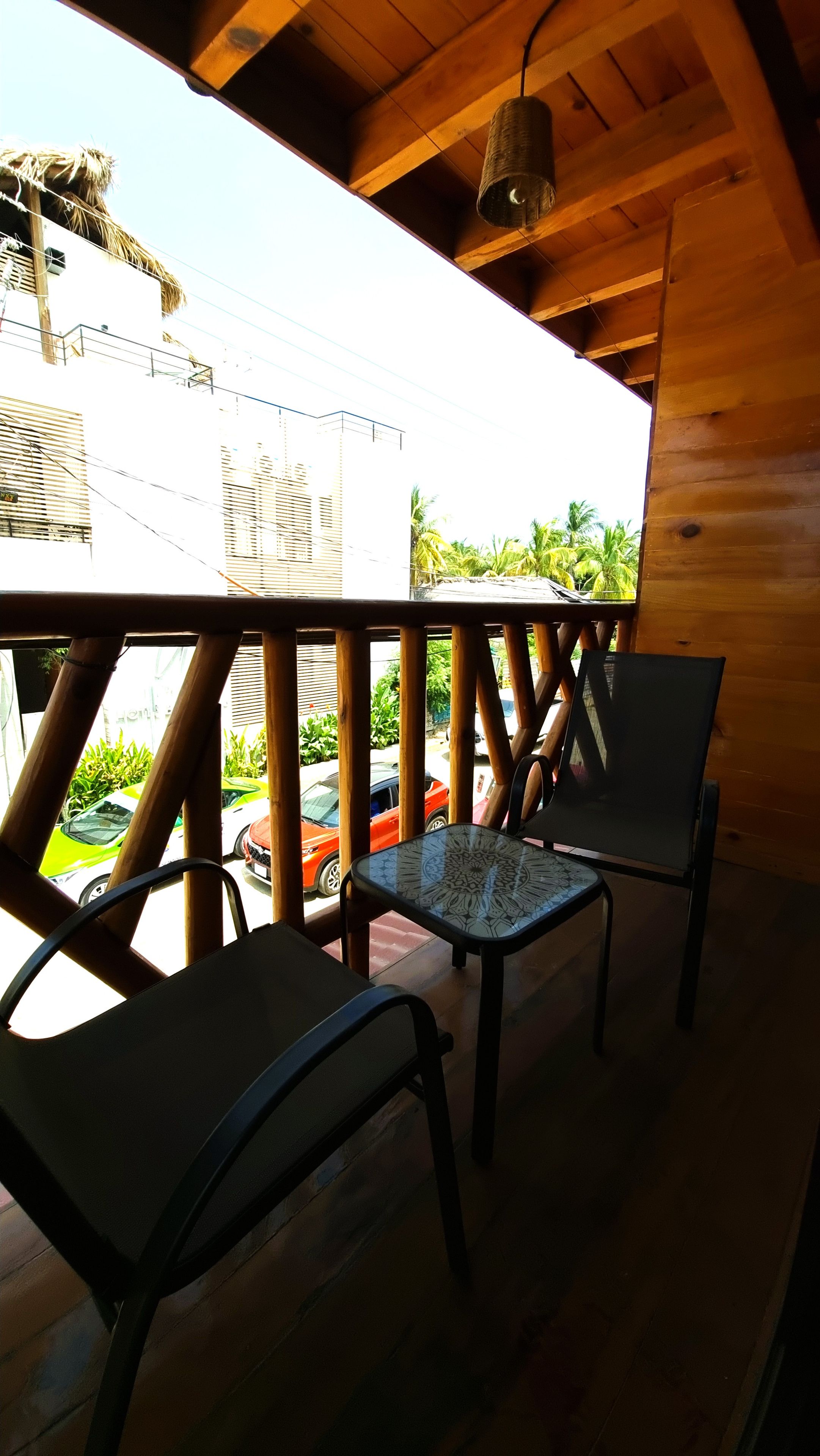 Una terraza de madera con dos sillas y una mesa, con vista a una calle con coches estacionados y vegetación.