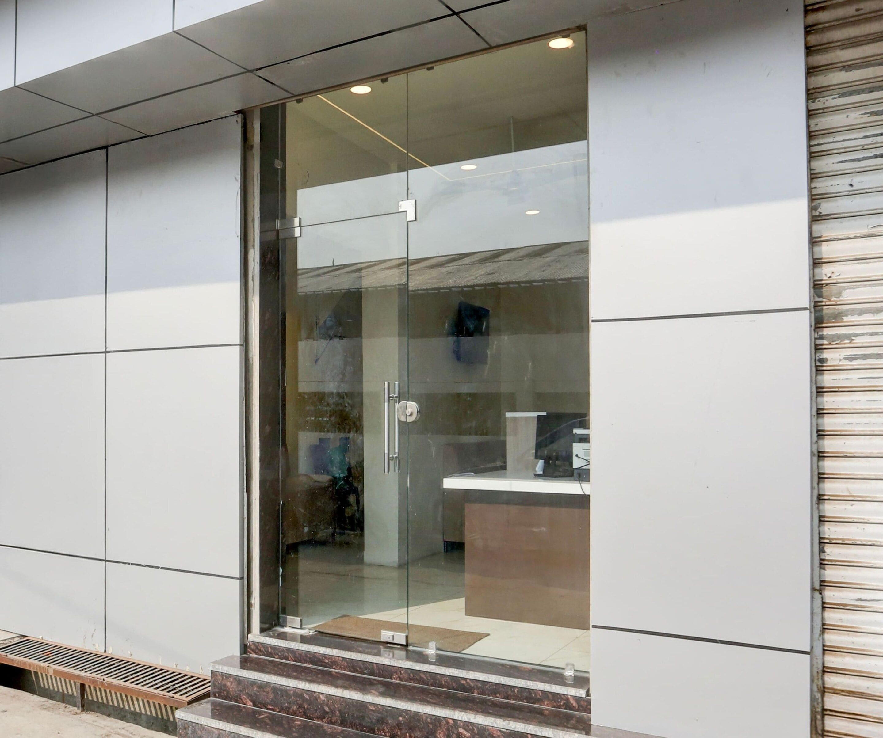 Una entrada moderna a un edificio con puerta de vidrio, escalera de metal y un mostrador de recepción en el interior.