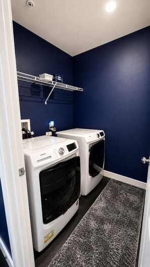 A laundry room with two front-loading washing machines, a drying rack, and a grey carpet. - Luxury 4BR | 3 King beds + Hot Tub + EV Charger (Victorville)