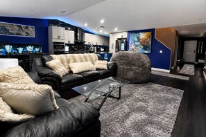 A modern living room with a black leather sofa, a glass coffee table, and a large gray shag rug. - Luxury 4BR | 3 King beds + Hot Tub + EV Charger (Victorville)