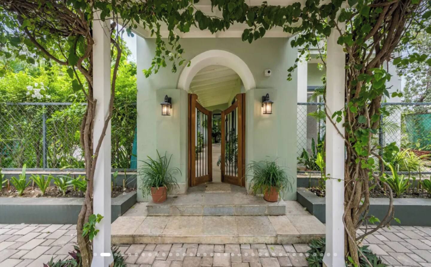 A covered entrance with a stone-paved pathway leading to a wooden door, flanked by potted plants and a metal fence.