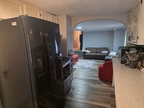 A compact kitchen with a black refrigerator, a built-in oven, and a microwave. There is a red chair and a grey sofa in the living area with a wooden door leading to another room. - Stunning 2-bedroom Unit with in-unit Laundry in Desirable Downtown Manchester (Manchester)