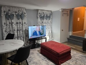 Un saló amb televisió que mostra un programa d'actualitat, taula de dinar rodona i un otoman vermell.