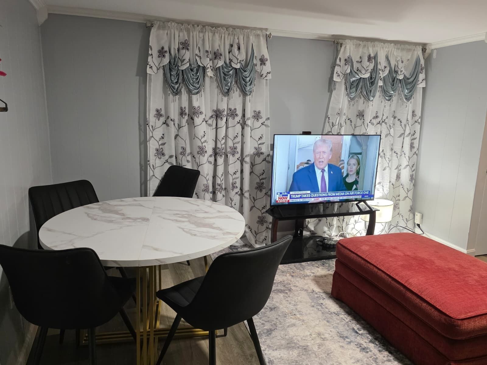 Una sala de estar con una mesa redonda, sillas negras, un reposapiés rojo y un televisor que muestra un noticiero.