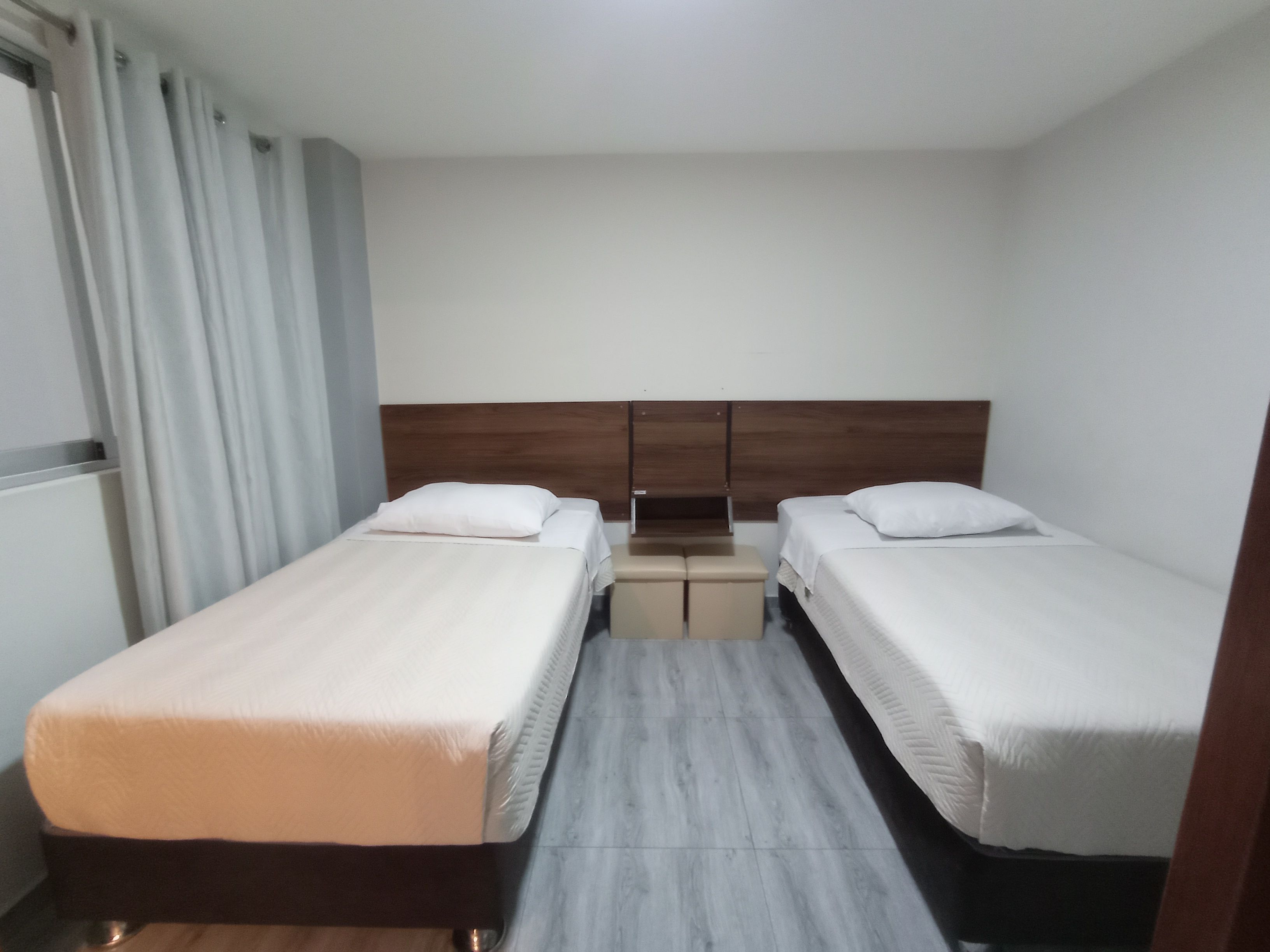 A hotel room with two single beds, a wooden headboard, a small table, and a window with curtains.