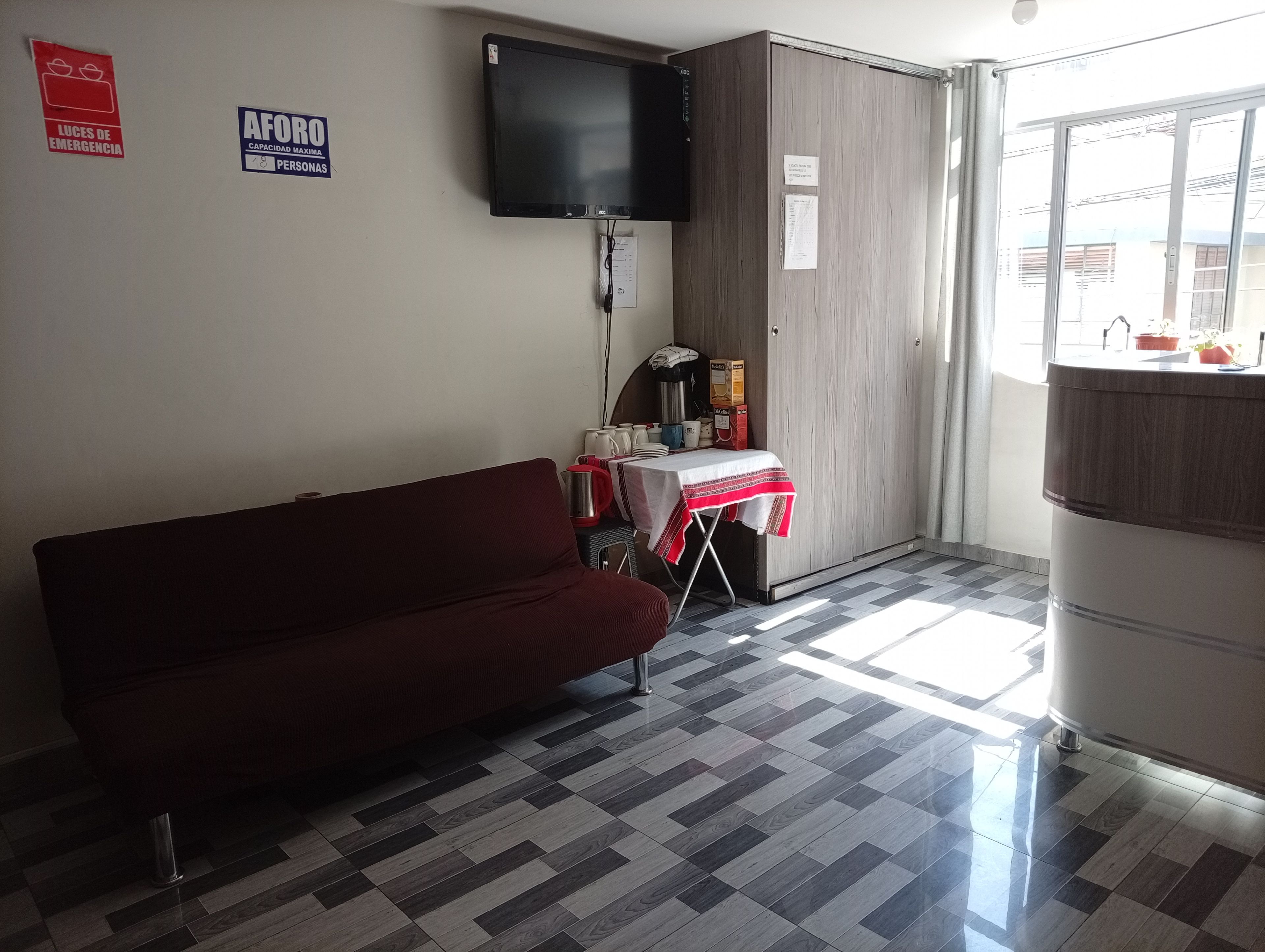 A room with a sofa, a table with a red and white tablecloth, a television mounted on the wall, and a window with a view of buildings.