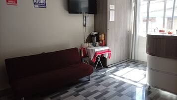 A room with a sofa, a table with a red and white tablecloth, a television mounted on the wall, and a window with a view of buildings.