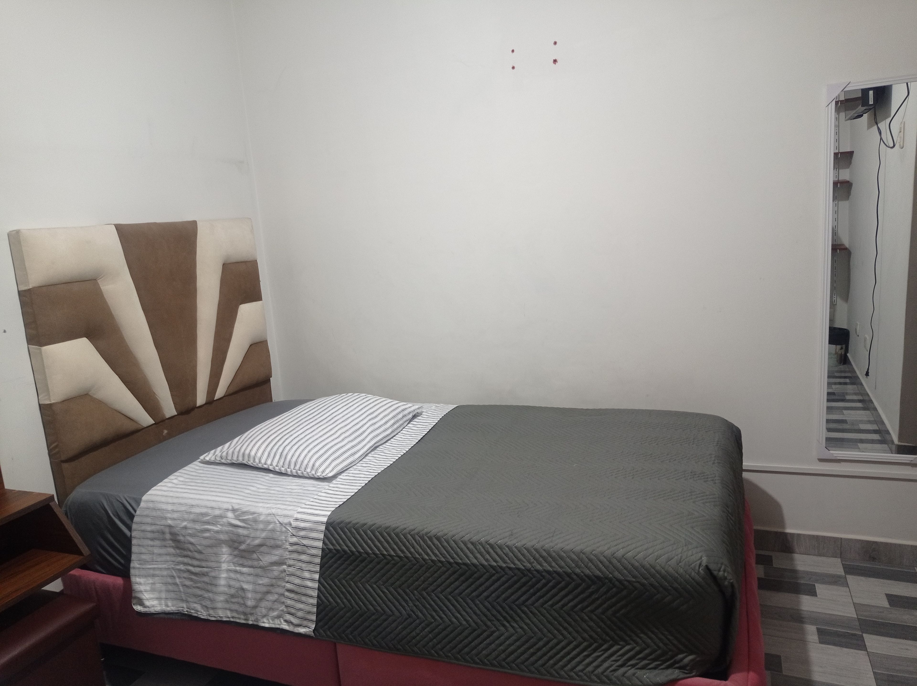 A single bed with a headboard, a bedside table, and a wall-mounted electrical outlet.