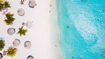 Una playa con agua turquesa cristalina, arena blanca y varias sombrillas de paja.