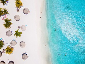 En strand med kristallklart turkosfärgat vatten, vit sand och flera parasoller med halmtak.