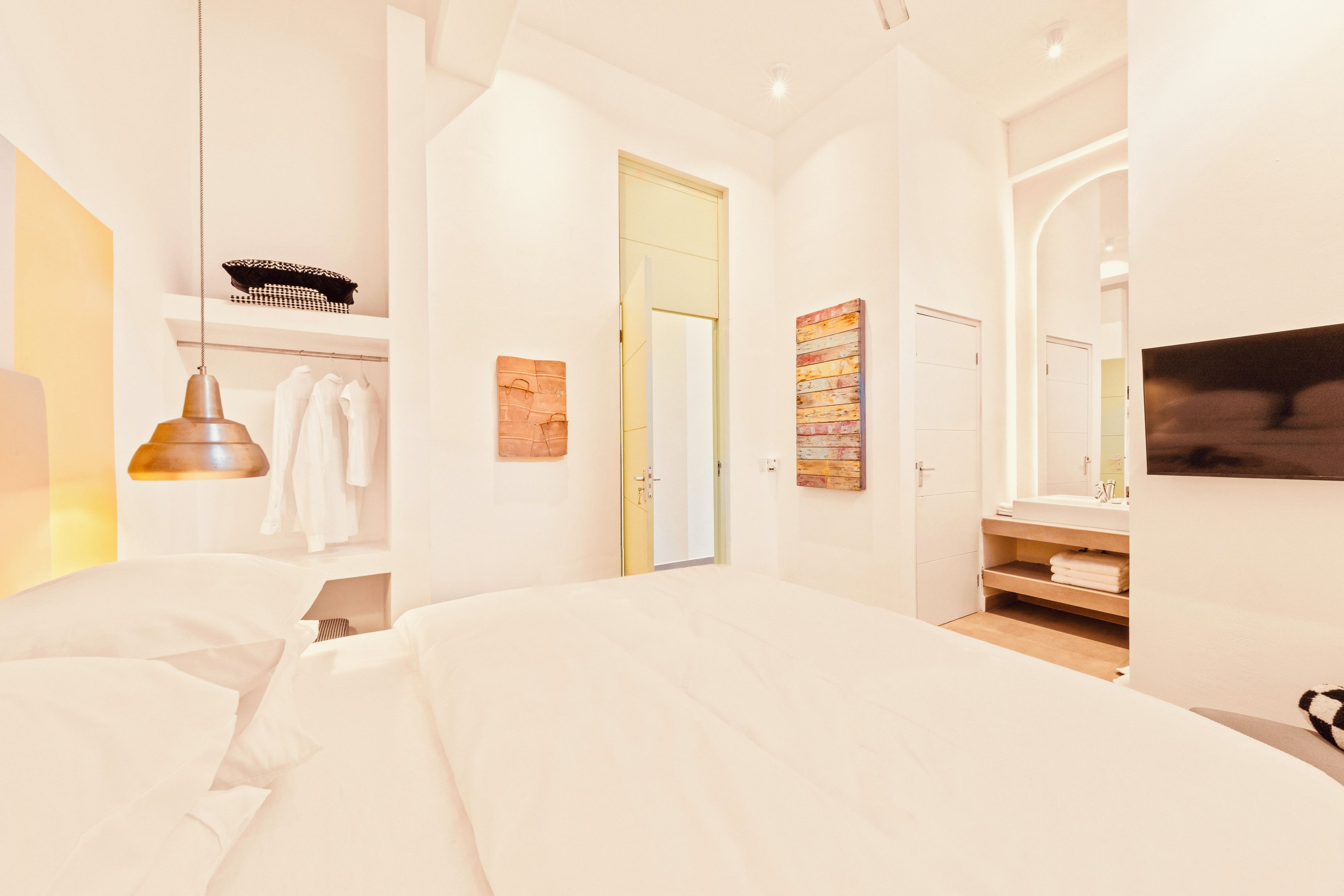 A modern bedroom with a large bed, a hanging light fixture, a wardrobe, and a bathroom with a mirror and sink.