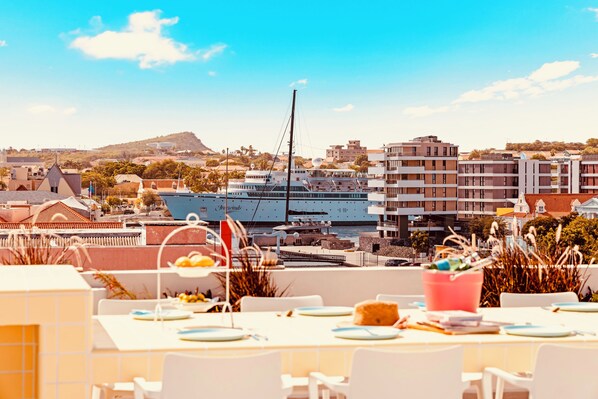 A dining area with tables set for guests, overlooking a marina with boats and buildings in the background.