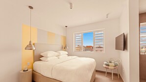 A bedroom with a bed, bedside lamps, a nightstand, a window with blinds, and a view of buildings outside. - BIKINI (Willemstad)