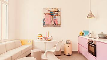 A modern kitchen with a pink cabinet, a white dining table, and a sofa with a yellow cushion.