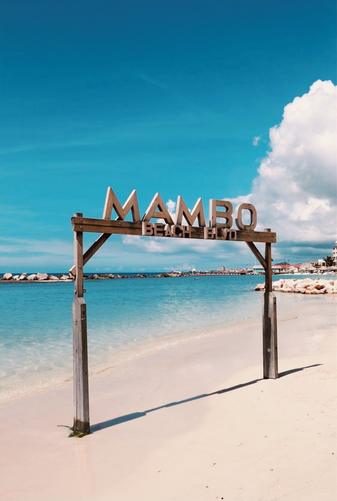 A wooden sign with the text 'MAMBO BEACH BLVD.' in front of a beach with clear blue water and a clear sky.