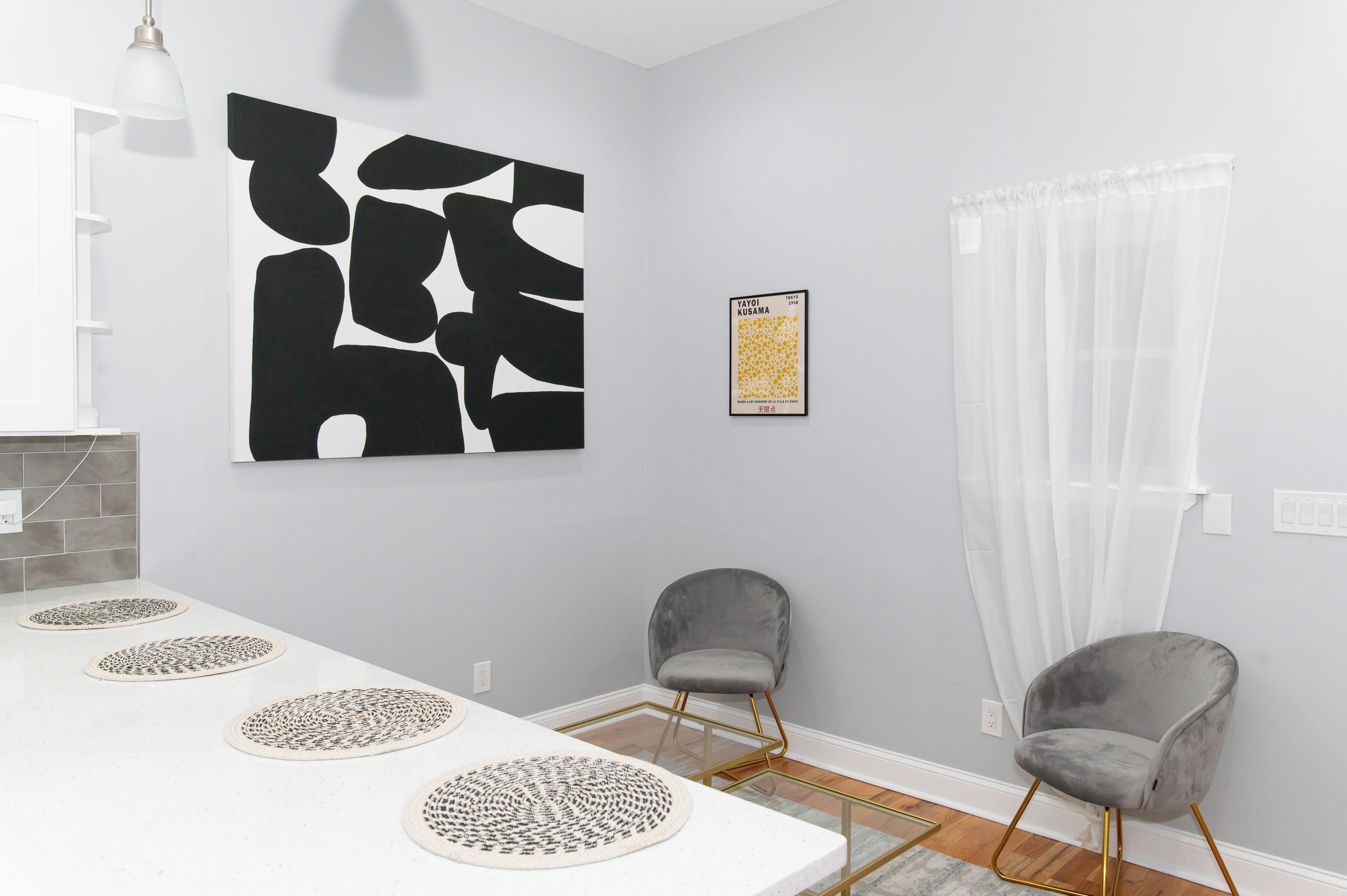 Un espace repas moderne avec une table blanche, deux chaises grises, un grand tableau abstrait, une œuvre d’art encadrée et un rideau blanc transparent.