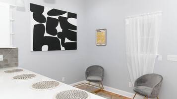 Uma sala de jantar moderna com uma mesa branca, duas cadeiras cinzentas, uma grande pintura abstrata, uma obra de arte emoldurada e uma cortina branca translúcida.