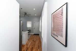 Un couloir avec un plancher en bois, un ventilateur de plafond, une porte blanche et une œuvre d’art encadrée accrochée au mur.